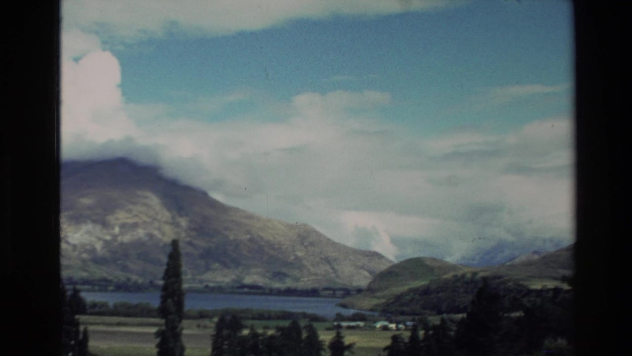 Mountain range beyond a lake with green fields and cloudy sky.