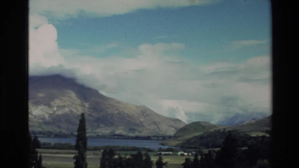 1982 New Zealand 8mm frame showing mountains, a lake, green valley ground, and bright clouds from a visitor viewpoint.