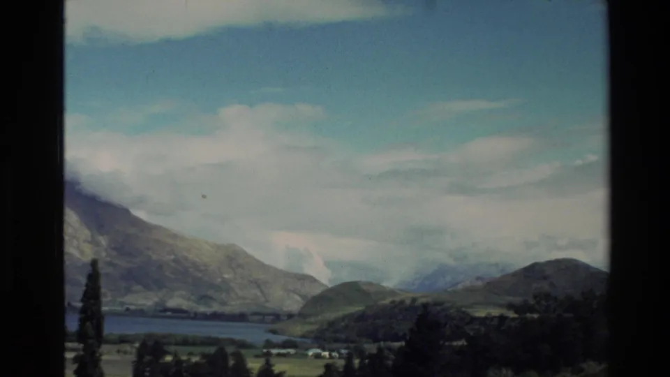 1982 New Zealand travel frame with a calm lake, layered hills, distant peaks, and bright cloud cover within visible 8mm frame edges.