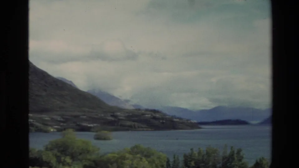 1982 New Zealand 8mm frame showing a broad lake, wooded foreground, sloping hills, and distant mountain silhouettes under overcast light.