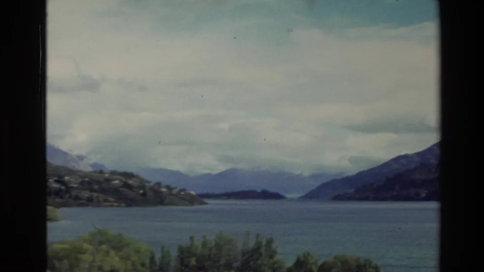1982 New Zealand home-movie frame showing lake water, a hillside with small light structures, distant mountains, and bright cloud cover.