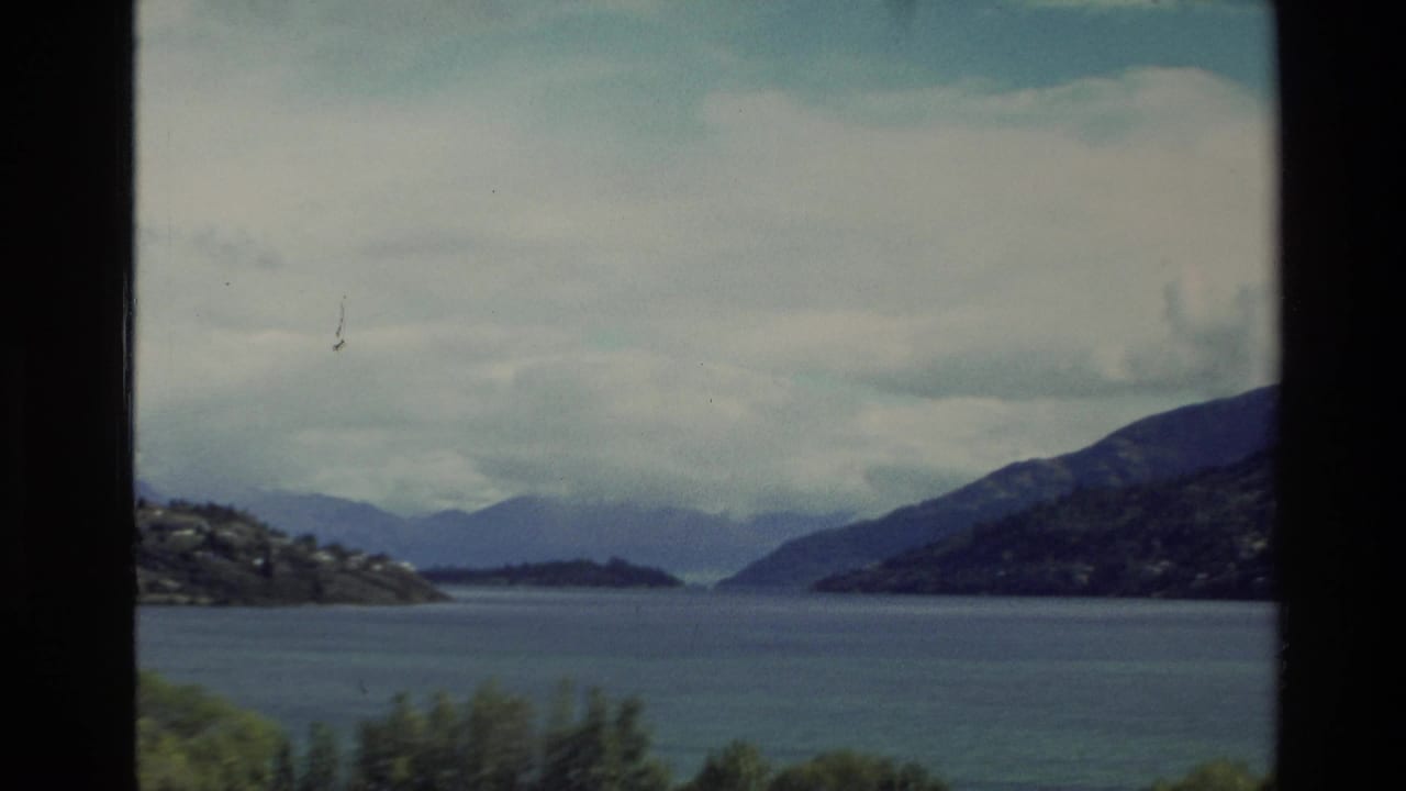 1982 New Zealand 8mm frame showing a blue lake, distant hills, shoreline trees, and heavy cloud cover.