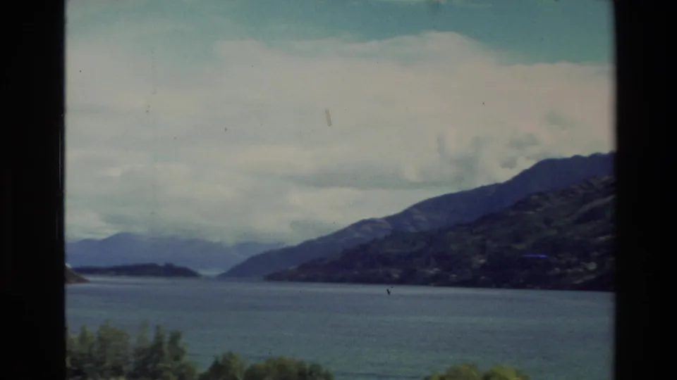 1982 New Zealand scenic frame with a calm blue lake, mountain shoreline, distant hills, and overcast sky.