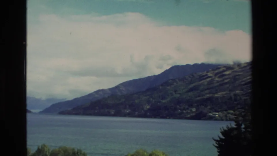 1982 New Zealand 8mm frame of a lake beside green hills, with shoreline trees and cloud-softened mountains.