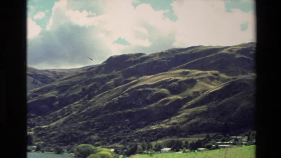 1982 New Zealand landscape frame showing rolling green mountains, a small valley settlement, trees, and bright clouds.