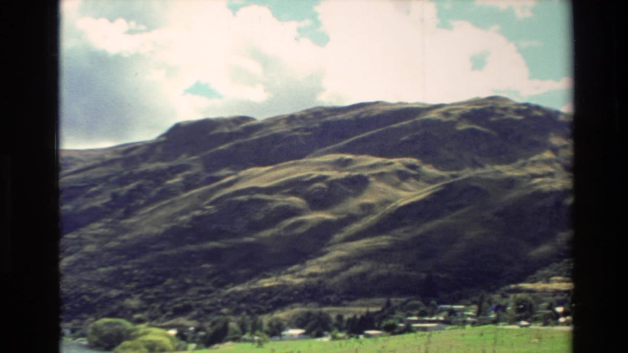1982 New Zealand 8mm frame showing green mountain slopes, scattered clouds, and rural buildings below the hills.