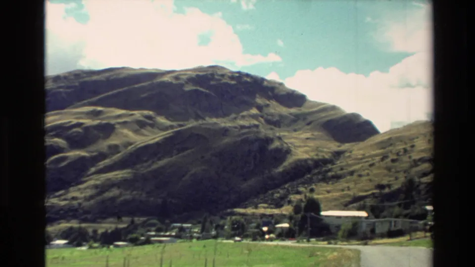 1982 New Zealand home-movie frame with rolling hills, a rural road, fields, and buildings below the slopes.