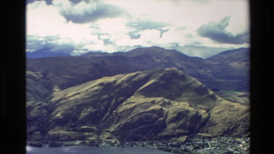 1982 New Zealand aerial home-movie frame showing sunlit hills, distant mountains, clouds, and a small lakeside or valley settlement.