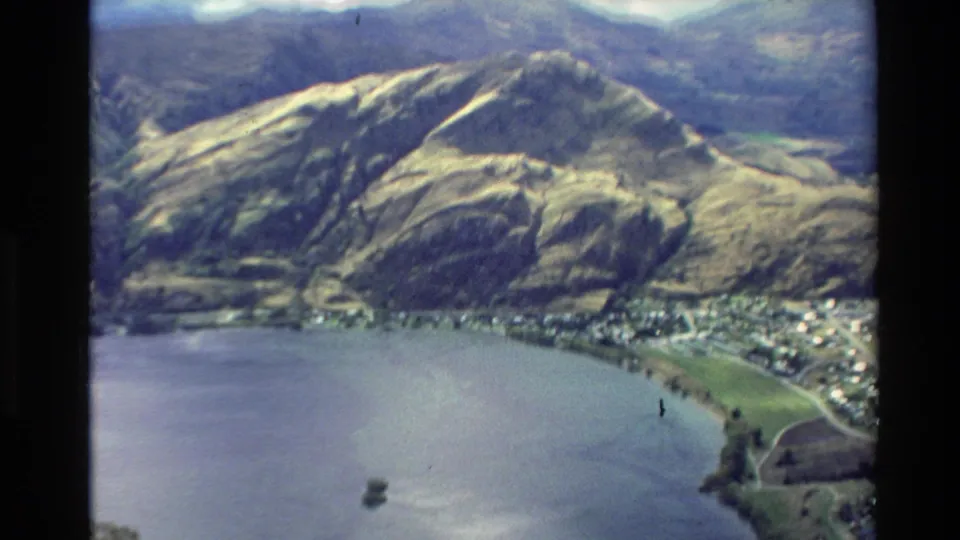1982 New Zealand 8mm aerial frame with a lake, shoreline town, fields, and steep rounded hills rising behind the water.