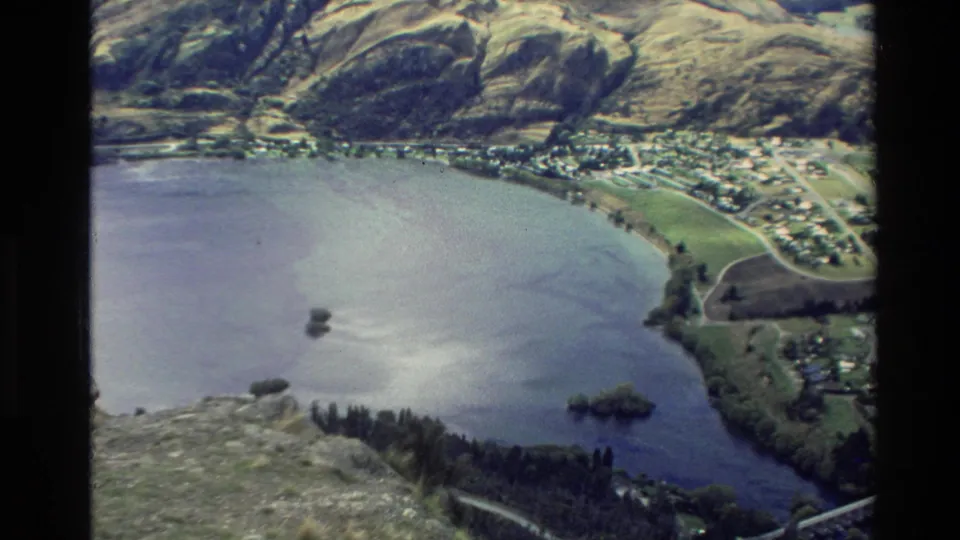 1982 New Zealand aerial view of a broad lake, wooded shoreline, small lakeside town, fields, and mountain slopes in soft 8mm color.