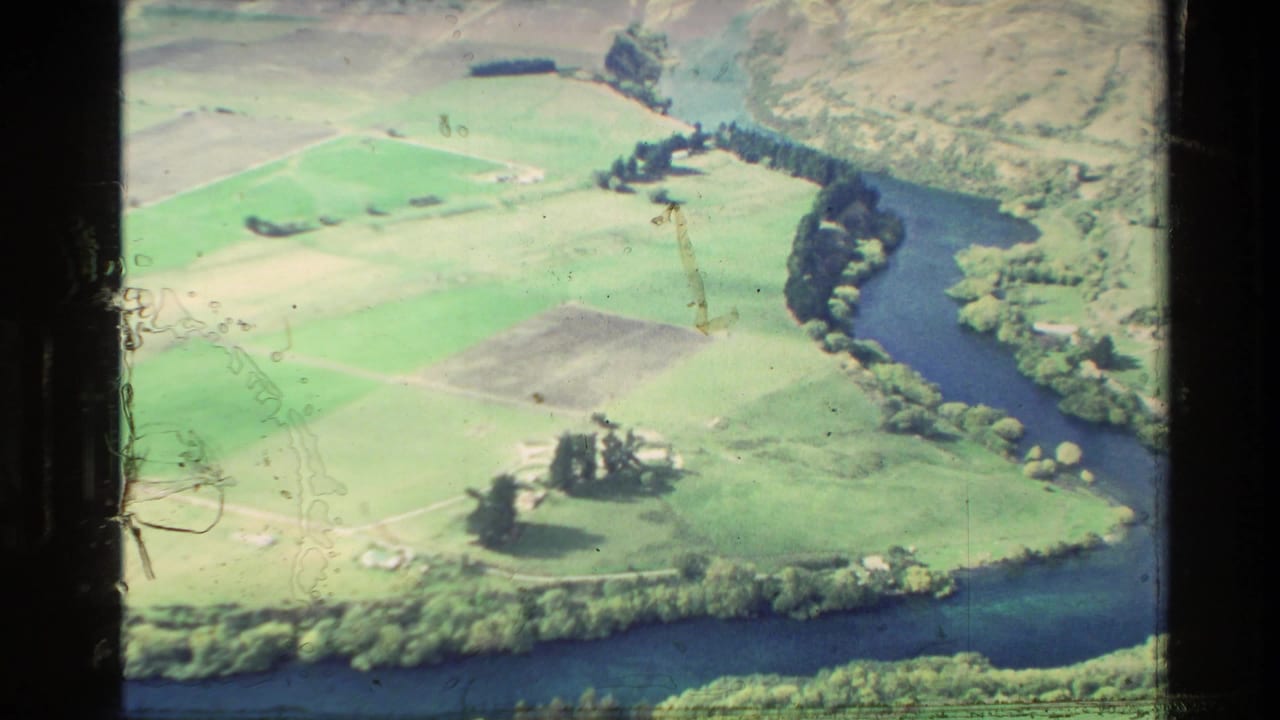 1982 New Zealand 8mm aerial view of green farmland, a curving river, tree belts, and small rural structures.