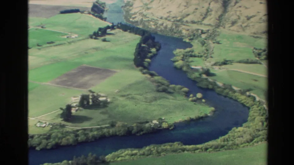 1982 New Zealand aerial frame showing a dark river bend, green paddocks, scattered trees, and hillside terrain.