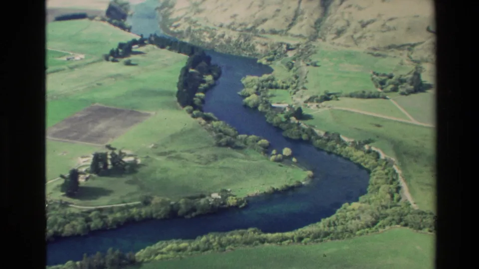 1982 New Zealand 8mm aerial view of a broad river curve, cultivated fields, trees, and rural tracks.