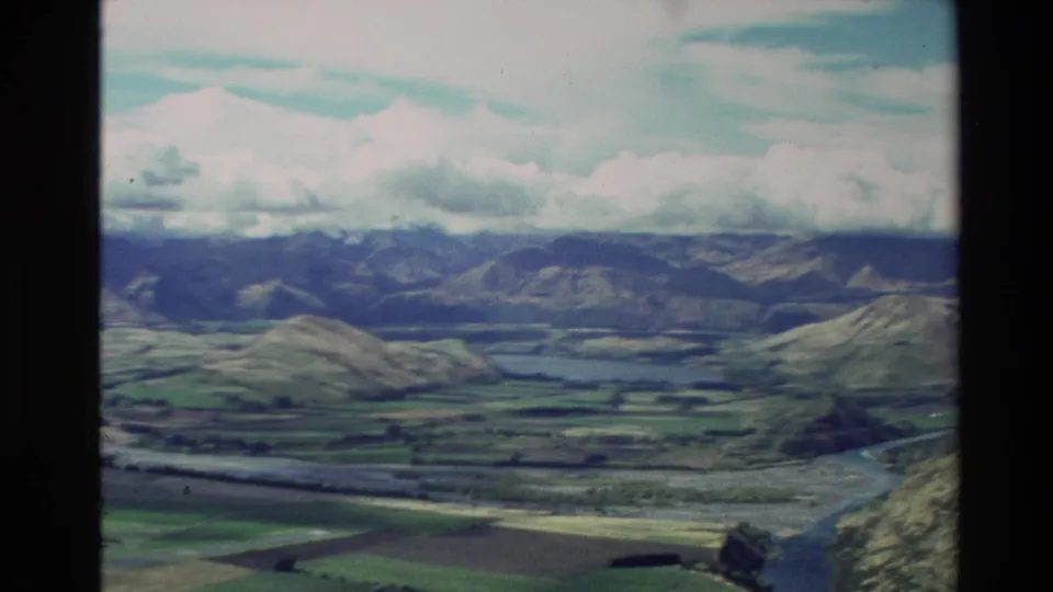 1982 New Zealand aerial landscape with a lake or wide waterway, patchwork fields, hills, and low clouds.