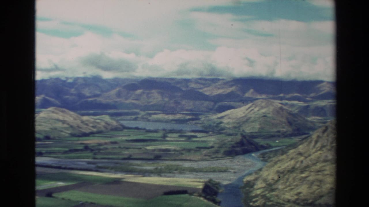 Aerial view over green hills, fields, a winding river, distant mountains, and clouded sky.