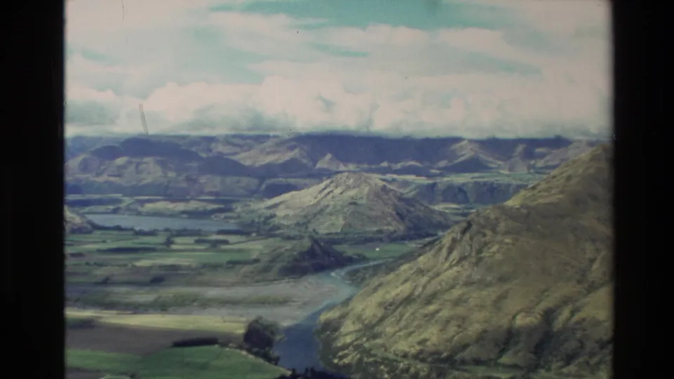 1982 New Zealand aerial frame with steep hills in the foreground, valley farmland, winding water, mountains, and cloud cover.