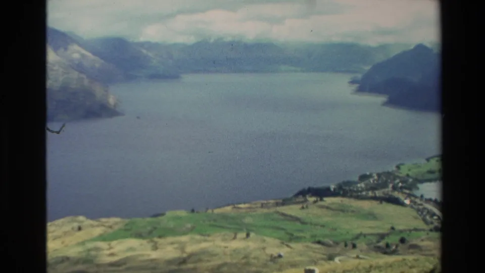 1982 New Zealand aerial home-movie view of a broad lake, mountain shoreline, green fields, and a small lakeside settlement.