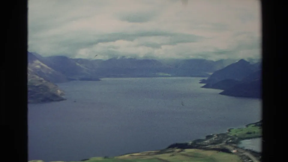 1982 New Zealand aerial frame showing a wide lake surrounded by mountain slopes, green shoreline patches, distant hills, and cloudy sky.