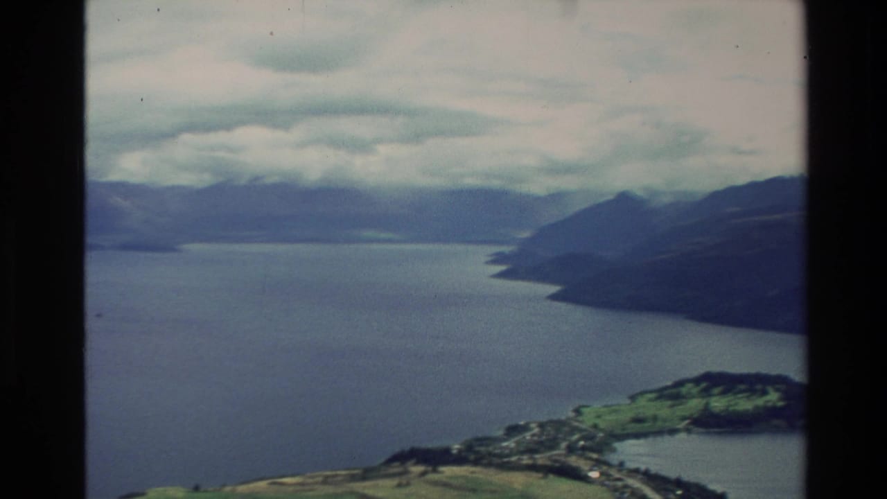 1982 New Zealand 8mm aerial view of a broad lake winding between dark mountain slopes and cloudy sky.