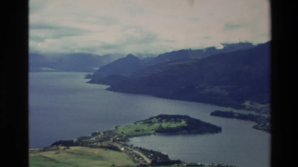 1982 New Zealand aerial frame showing a lake valley, mountain ridges, green shoreline fields, and a small settlement.