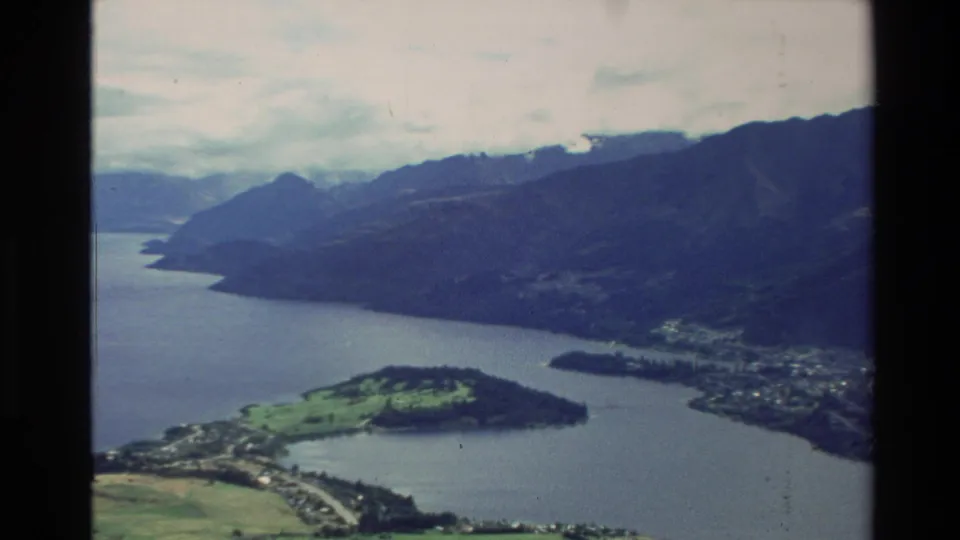 1982 New Zealand 8mm frame of a lake bordered by mountains, with a green peninsula, roads, and shoreline buildings below.