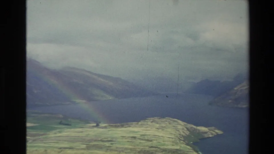1982 New Zealand aerial frame showing a misty lake valley, mountain slopes, open hillside, and a faint rainbow.