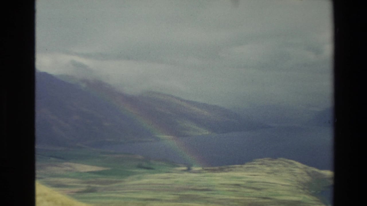 1982 New Zealand 8mm frame showing a faint rainbow over misty mountains, a lake, and rolling grassland.