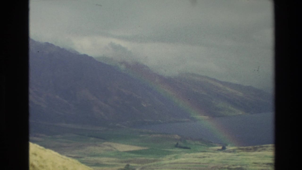 Rainbow over dark mountain slopes, water, and open grassland under cloud.