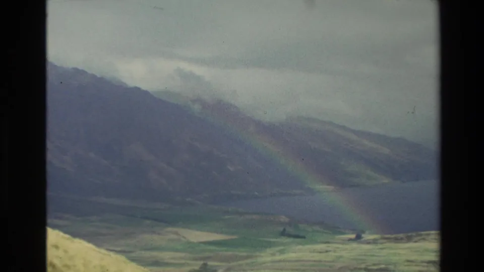 1982 New Zealand 8mm frame with a rainbow descending across dark mountain slopes beside a lake and grassy valley.
