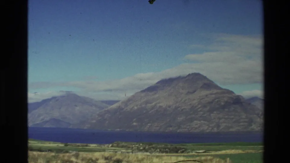 1982 New Zealand 8mm frame showing clearer skies over rugged mountains, deep blue water, and green foreground land.