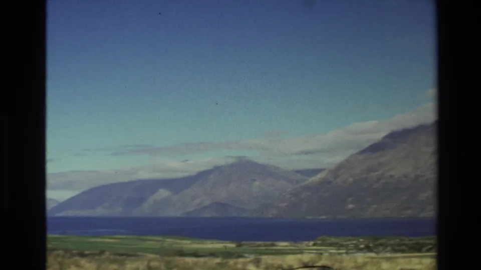 1982 New Zealand 8mm frame with bright blue sky, distant mountain ridges, and a dark blue lake or inlet.