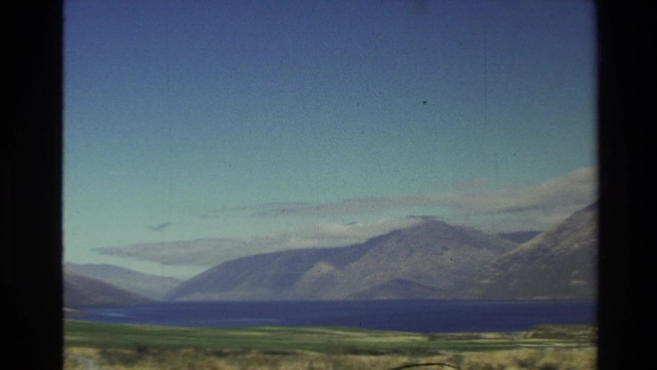 1982 New Zealand home-movie frame showing a blue lake below rugged mountains, filmed from a moving roadside viewpoint.