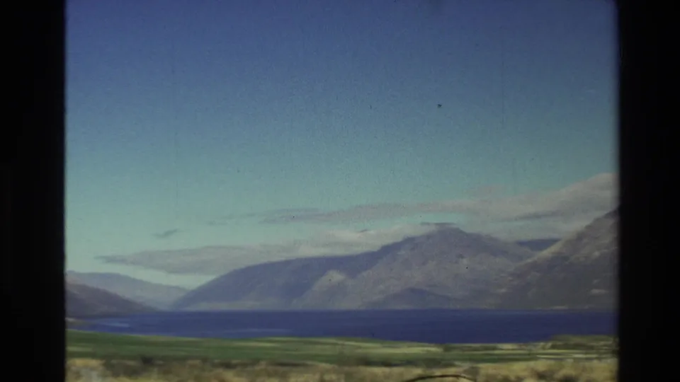 1982 New Zealand home-movie frame showing a blue lake below rugged mountains, filmed from a moving roadside viewpoint.
