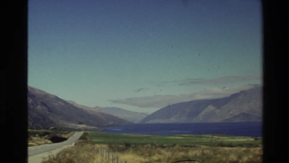 A New Zealand lakeside road curves through open grassland toward mountains in this 1982 8mm travel frame.