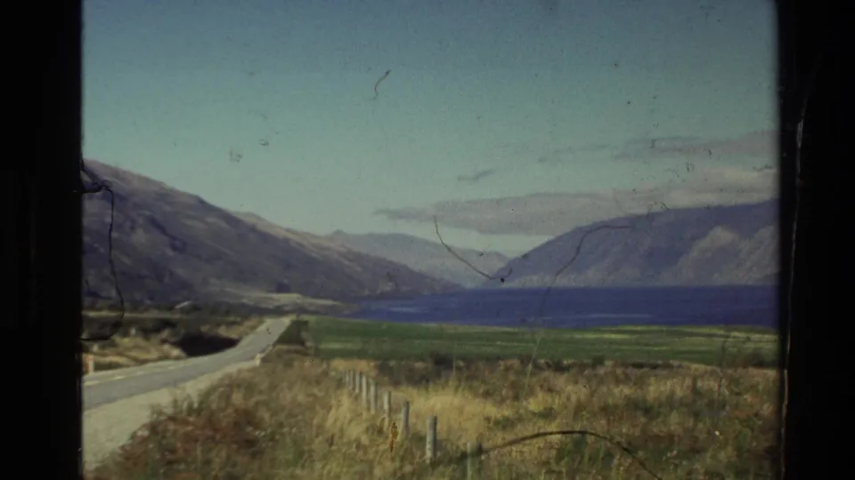 Moving-vehicle 8mm frame of a New Zealand shoreline road, grassy verge, lake water, and mountains under a pale sky.