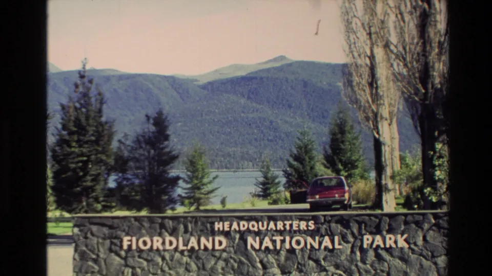 Fiordland National Park headquarters sign in New Zealand, with a red car, tall trees, lake water, and wooded mountains beyond.