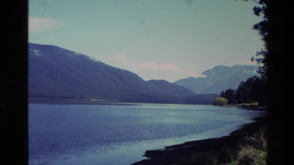 Wide 1982 New Zealand lake view with dark mountain slopes, distant peaks, shoreline vegetation and pale sky.