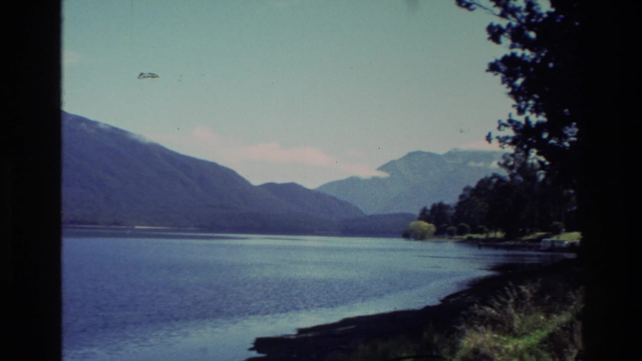 1982 New Zealand 8mm frame showing a mountain lake, wooded shoreline, distant peaks, and dark trees at the edge of the view.