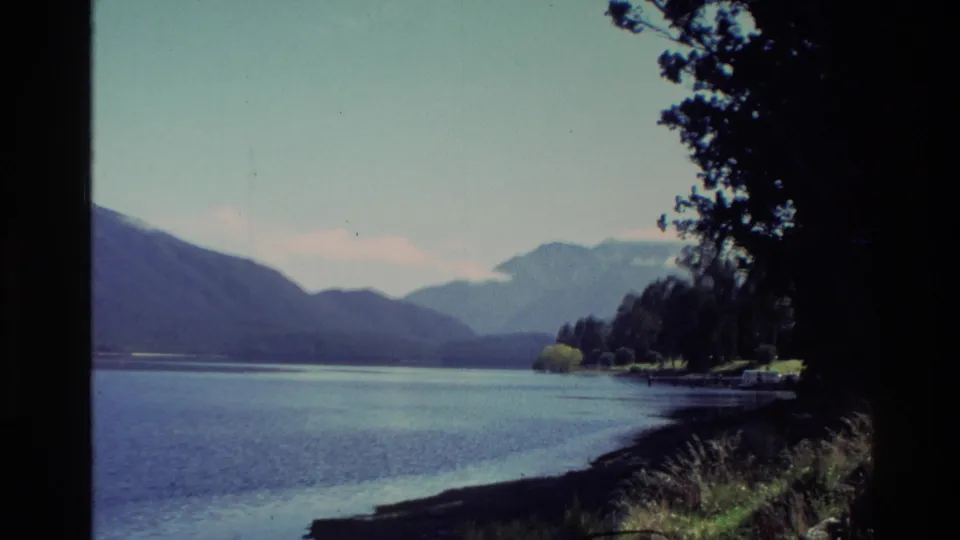 1982 New Zealand 8mm frame of a calm lake with a tree-lined shore, mountain ridges, and foreground tree silhouettes.