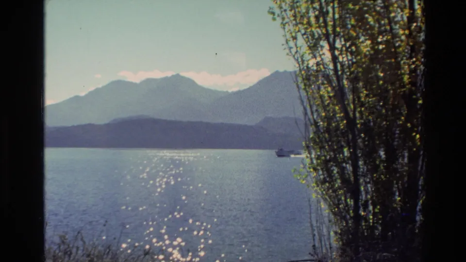 1982 New Zealand 8mm frame showing glittering lake reflections, distant mountains, shoreline trees, and a small boat crossing the water.