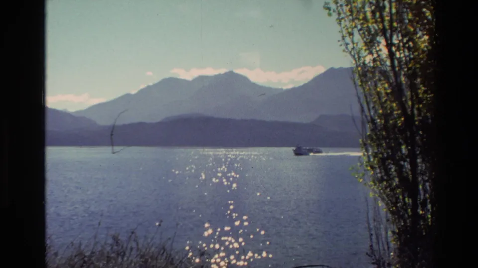 1982 New Zealand 8mm frame showing a motorboat on a calm lake, with sparkling water, distant mountains, and dark foliage at the edge.