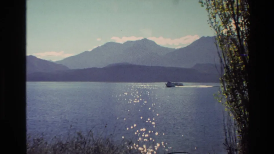 1982 New Zealand 8mm frame of a small motorboat skimming across open lake water, backed by hazy mountains and a pale sky.