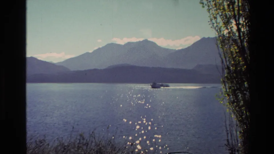 1982 New Zealand 8mm frame with a small motorboat on a calm lake, dark foreground grasses, a tree line at right, and mountains beyond.
