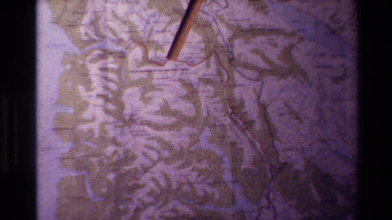 1982 New Zealand home-movie frame showing a close-up of a paper topographic map, red route line, and wooden pointer under soft 8mm color shift.
