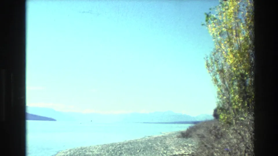 New Zealand lakeside view from 1982 8mm footage, with bright blue water, a rocky shore, distant mountains, and tall trees along the right side.