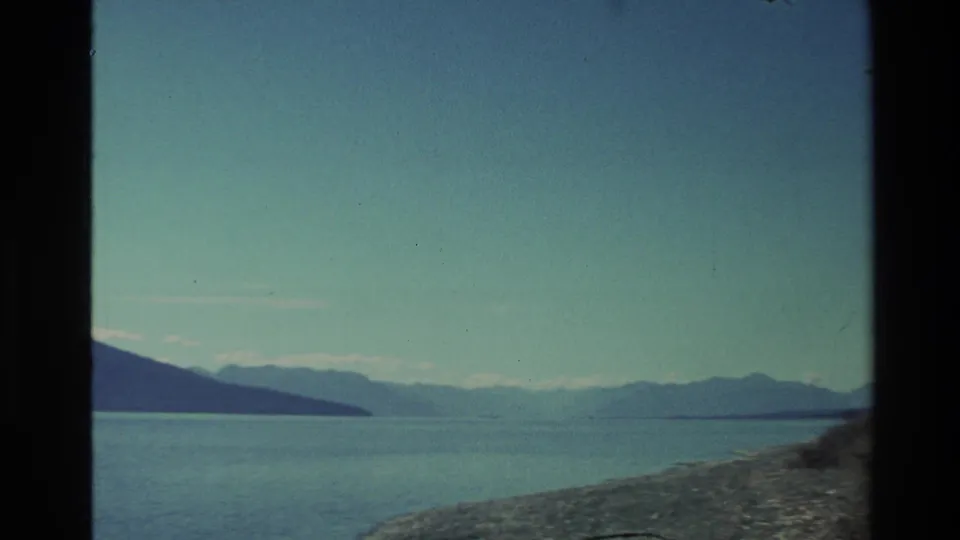 Scenic New Zealand lake landscape on 8mm film, showing calm water, layered distant mountains, rocky shoreline, and clear sky.