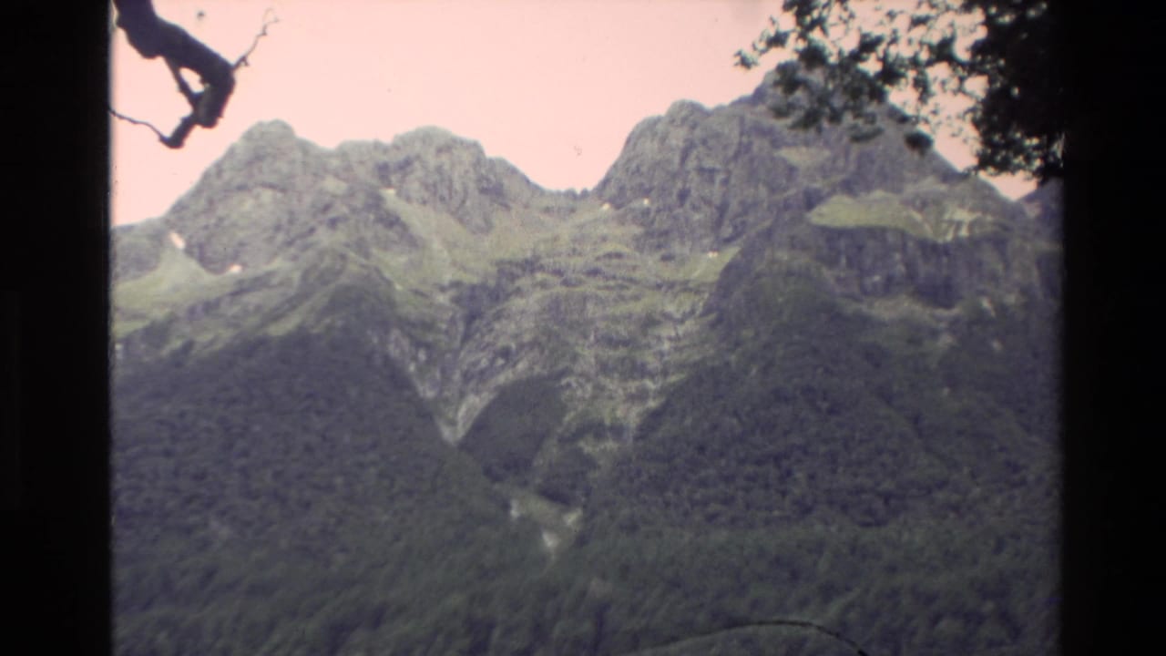 1982 New Zealand 8mm frame showing rugged mountain ridges above a dense forested valley, with dark film borders and a pale sky.