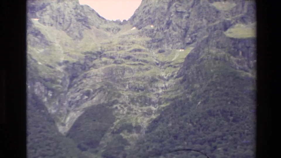 1982 New Zealand 8mm frame showing a closer section of craggy mountain terrain, green ledges, and dark forested valley below.