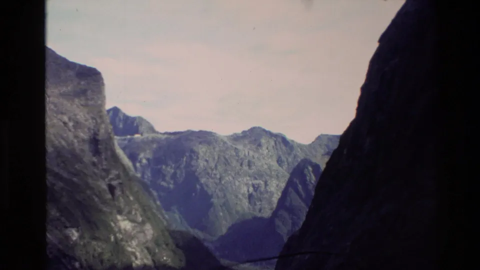 1982 New Zealand 8mm frame with a dark cliff edge, green valley walls, and layered mountain ridges in soft color.
