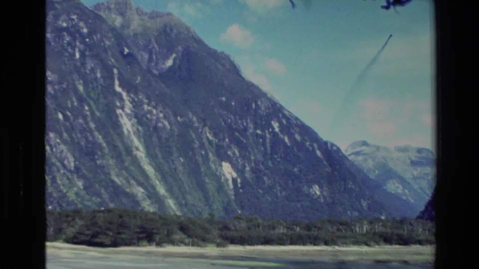 1982 New Zealand 8mm frame showing a steep rocky mountain, forested shoreline, open water, and distant snowy peaks under blue sky.